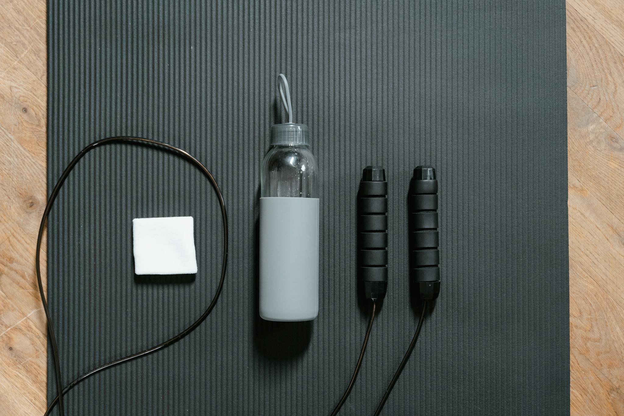 Flatlay of jump rope, water bottle, and towel on a black yoga mat.